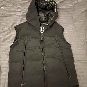 Säljer en snygg svart dunväst från Moncler i mycket bra skick. Västen har en dragkedja framtill och en cool logga på insidan av huvan. Perfekt för höst och vår när du vill hålla dig varm men ändå se stilren ut. Passar både till vardags och fest! Storlek 1 ungefär S-M