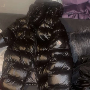 Svart dunjacka från Moncler.  - Säljer en snygg och varm svart dunjacka från Moncler. Jackan har en glansig finish och är perfekt för kalla vinterdagar. Den har en dragkedja framtill och en klassisk Moncler-logga på ärmen. Insidan har en cool serietidningsdesign. Den är äkta och har även nfc Scan.det är storlek M men passar S med 