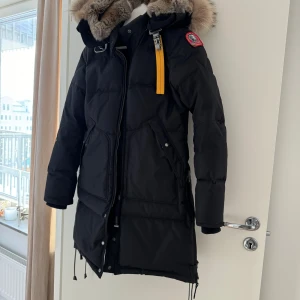 Parajumper Svart dunjacka med pälsdetaljer - Säljer en svart Parajumpers dunjacka i mycket bra skick. Den har en mysig pälsfodrad huva och en cool gul dragkedjedetalj. Perfekt för kalla vinterdagar! Jackan har långa ärmar och flera praktiska fickor. Passar både till vardags och mer äventyrliga utflykter.