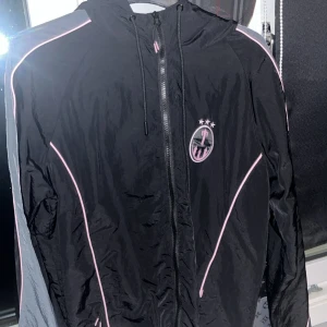 Svart vindjacka från Trapstar och byxor  - Säljer en svart vindjacka från Trapstar med rosa detaljer och dragkedja. Jackan har en cool logga på bröstet och är perfekt för vår och höst. Den är i mycket bra skick och har en normal passform. Perfekt för streetwear-stilen!