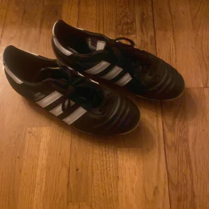 Adidas fotbollsskor i svart och vitt - Säljer ett par klassiska Adidas fotbollsskor i svart och vitt. Skorna har snörning och är i bra skick med tydliga dobbar för bra grepp på planen. Perfekta för fotbollsträning eller match. Tillverkade i Tyskland och redo för nya äventyr på fotbollsplanen!