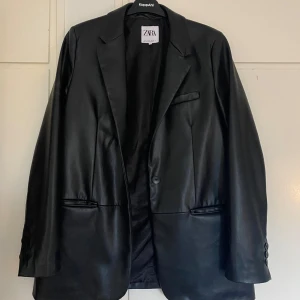 Svart kavaj från Zara - Snygg svart kavaj från Zara i skinnimitation. Perfekt för både vardag och fest! Den har en klassisk design med knappar fram och fickor på sidorna. Passar bra till höst och vår. Ett måste i garderoben för en stilren look.
