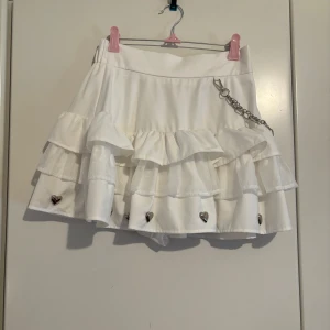 Ank Rouge Vit volangkjol med hjärtan och kedjedetalj - Superfin vit kjol med volanger och söta hjärtan längst ner. Den har en cool kedjedetalj som ger en edgy touch. Kjolen är kort med inbyggda shorts och har en dragkedja bak för enkel påklädning. Kjolen är från det japanska märket Ank Rouge! 