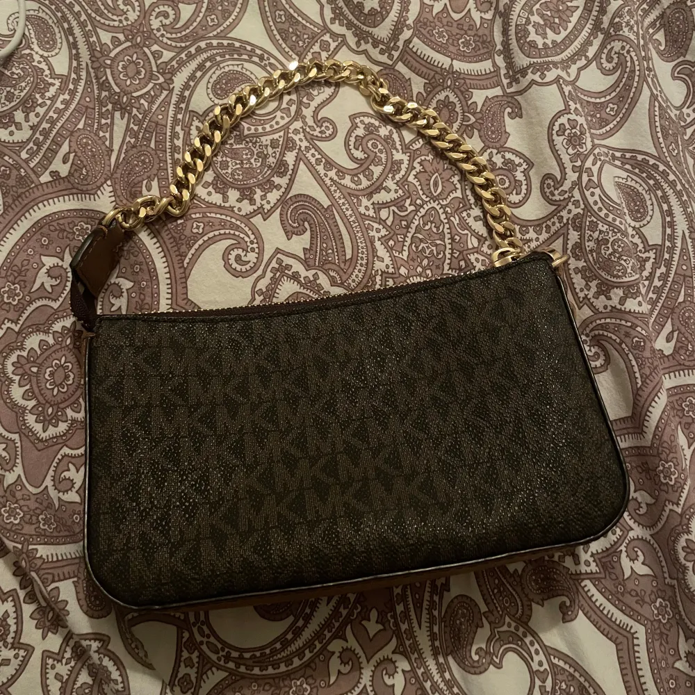 Snygg brun axelväska från Michael Kors med guldfärgad kedja och dragkedja. Väskan har ett diskret mönster och är perfekt för både vardag och fest. Den är i mycket bra skick och redo att användas direkt. Perfekt för att ge din outfit en lyxig touch!. Väskor.