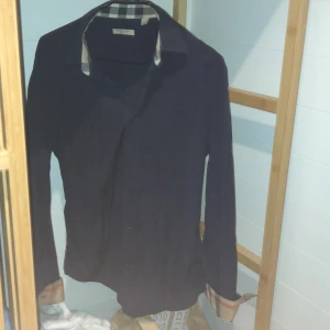 Svart skjorta från Burberry - Säljer en stilren svart skjorta från Burberry i bra skick. Den har ett klassiskt rutigt mönster på insidan av kragen och knappar framtill. Perfekt för både vardag och fest! 🖤En knapp har gått av men som inte syns för färgen är marinblå pris kan diskuteras 