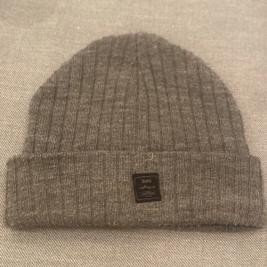 Grå ribbad mössa från Barts - Säljer en snygg grå ribbad mössa från Barts. Perfekt för kalla dagar med sin mjuka och varma känsla. Den har en uppvikt kant och en liten etikett framtill. Passar till alla vinteroutfits!