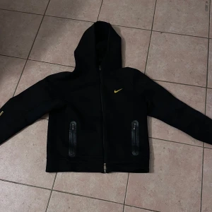 Svart huvtröja från NOCTA - Säljer en svart huvtröja från NOCTA i mycket bra skick. Den har en dragkedja framtill och två fickor med dragkedjor. Tröjan har en gul Nike-logga på bröstet och små gula detaljer på baksidan. Perfekt för höst och vår!