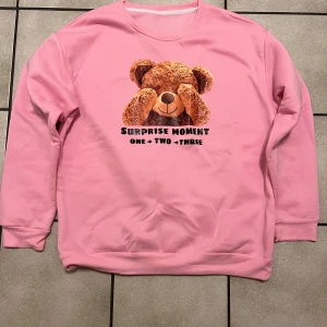 Rosa sweatshirt med nallemotiv - Supergullig rosa sweatshirt med ett stort nallemotiv på framsidan och texten 'Surprise Moment One Two Three'. Perfekt för mysiga dagar eller som en söt present. Tröjan har långa ärmar och en bekväm passform.