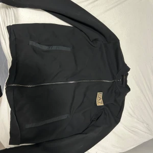 Armani tröja  - Säljer en stilren svart tröja från EA7 Emporio Armani. Den har en dragkedja framtill och en snygg logga på bröstet. Perfekt för höst och vår, med en normal passform och långa ärmar. Jackan är i mycket bra skick och men passar bara inte mig helt enkelt. Lite stor för mig. Köpt för 1300kr