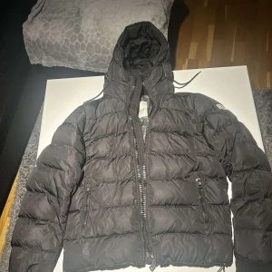 Svart dunjacka moncler  - Säljer en snygg svart dunjacka från Moncler i mycket bra skick. Jackan har en quiltad design med dragkedja och huva, perfekt för kalla dagar. Den är långärmad och har en normal passform. Moncler-loggan syns på ärmen. Perfekt för höst och vinter!