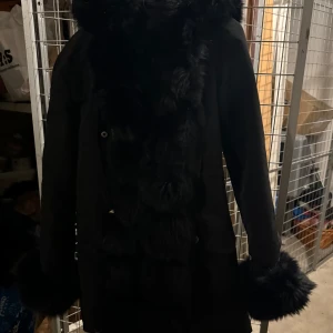 Svart kappa med pälsdetaljer - Säljer denna fina populära jacka! Kappa med fluffiga pälsdetaljer vid armar och luva även längs framsidan. Jackan är ock väldigt varm! Köpte denna för flera år sedan men efter Google sökningar ligger denna på 3500 kr och säljer det för 2500 kr. Tyckte det var svårt att få en ärlig bild på denna och därför tog jag även med en från en hemsida. Detta är fotat snabbt vid mitt förråd:) 