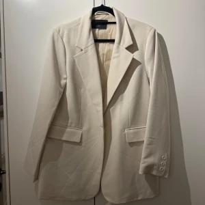 Krämvit kavaj från Bik Bok - Säljer en stilren beige kavaj från Bik Bok i storlek XS/S. Kavajen har en klassisk design med knappar på ärmsluten och fickdetaljer fram. Perfekt för både vardag och festliga tillfällen. Passar bra till höst och vår!