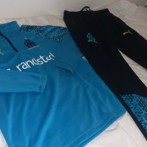 Blå marseille tracksuit - Säljer en blå marseille träningsset från Puma med långärmad tröja och byxor. Tröjan har ett coolt mönster på axlarna och byxorna har en matchande detalj vid bensluten. Perfekt för träning eller chill!
