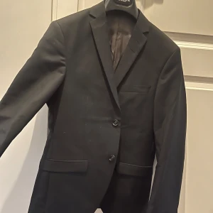 Svart kostym från Tiger of Sweden - Säljer en elegant svart kostym från Tiger of Sweden i ull och mohair. Kavajen har två knappar och en klassisk passform. Perfekt för formella tillfällen eller en stilren look på jobbet. Byxorna är matchande och ger en komplett outfit. Kvalitetstyg från Vitale Barberis Canonico.