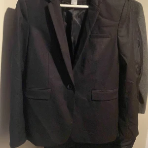 Svart kavaj från H&M - Snygg svart kavaj från H&M i storlek 40. Perfekt för både jobb och festliga tillfällen. Kavajen har en klassisk design med en knapp framtill och två fickor. Materialet känns bekvämt och ger en stilren look. Ett måste i garderoben för en elegant touch!