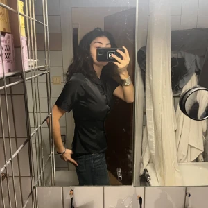 Svart kortärmad blus - Snygg svart blus i satin med korta ärmar och knappar framtill. Perfekt för både fest och vardag. Den har en figurnära passform som ger en elegant look. Passar bra till jeans eller kjol för en stilren outfit.