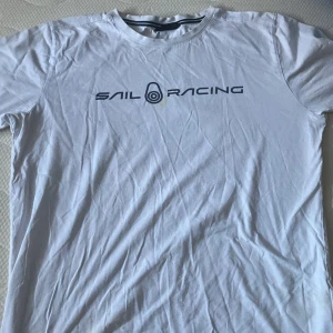 Vit t-shirt från Sail Racing - Säljer en vit t-shirt från Sail Racing i bra skick. Den har en stilren design med logotypen tryckt på framsidan och ett litet märke på baksidan. Perfekt för en avslappnad stil eller sommarens äventyr. Passar både till vardags och till träning. Har ett litet håll och en liten fläck som jag försökt tvätta bort men det går inte!🙂