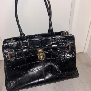 Svart handväska med krokodilmönster - Snygg svart handväska från Calvin klein i skinn med krokodilmönster och gulddetaljer. Väskan har ett elegant lås framtill och flera fack inuti för bra organisering. Perfekt för både vardag och fest👩‍❤️‍💋‍👩