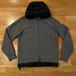 Grå stickad jacka med huva från Moncler - Säljer en snygg grå stickad jacka från Moncler med svart quiltad huva. Perfekt för höst, vår och svensk vinter! Den har en dragkedja framtill och är i mycket bra skick 10/10. Jackan är långärmad och har en normal passform M/L. Hör av er vid intresse eller frågor!                                                       