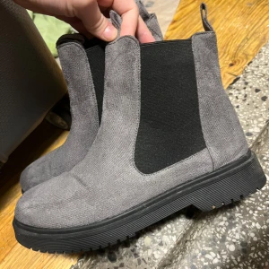 Grå Chelsea boots i mocka - Snygga grå Chelsea boots i mocka med elastiska svarta paneler på sidorna för enkel på- och avtagning. De har en robust svart sula som ger bra grepp. Perfekta för höst och vår, dessa boots är både stiliga och bekväma. Passar till både vardag och lite finare tillfällen. 