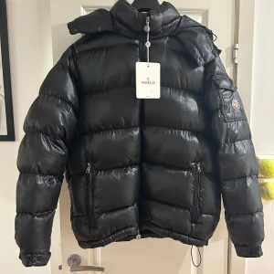 Svart dunjacka från Moncler Maja - Säljer en snygg svart dunjacka från Moncler i nytt skick. Jackan har en pufferstil med långärmade ärmar och dragkedja framtill. Den är perfekt för vintern och håller dig varm och stilren. Moncler-loggan syns på ärmen och den har en avtagbar huva. Passar perfekt för kalla dagar!