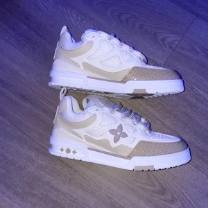 Beige och vita sneakers från Louis Vuitton - Säljer ett par snygga Louis Vuitton sneakers i beige och vitt. De har en stilren design med LV-logga på sidan och sulan. Skorna är i syntetmaterial med snörning och kommer i originalpåse. Perfekta för både vardag och fest!