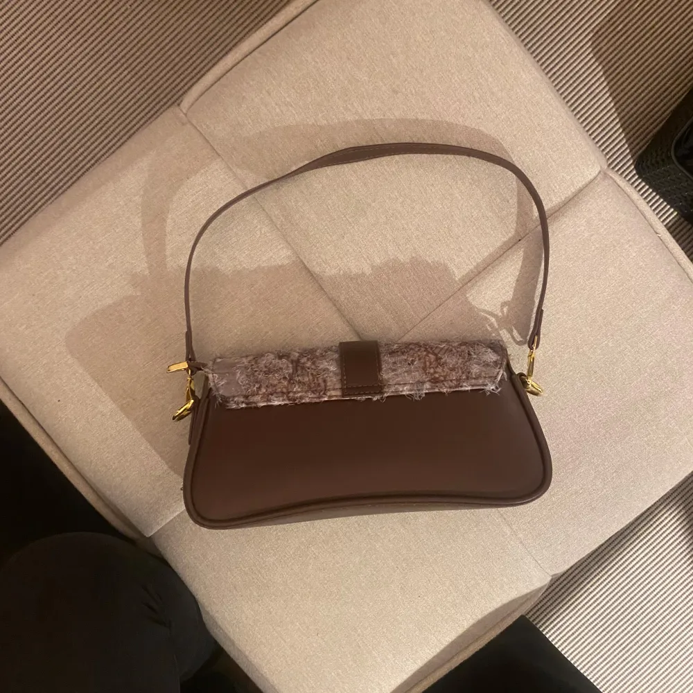 Snygg brun axelväska med unik mönstrad framsida och gulddetaljer. Väskan har en praktisk dragkedja och en stilren design som passar perfekt för både vardag och fest. Den är i mycket bra skick och redo att användas direkt!. Laukut & Käsilaukut.