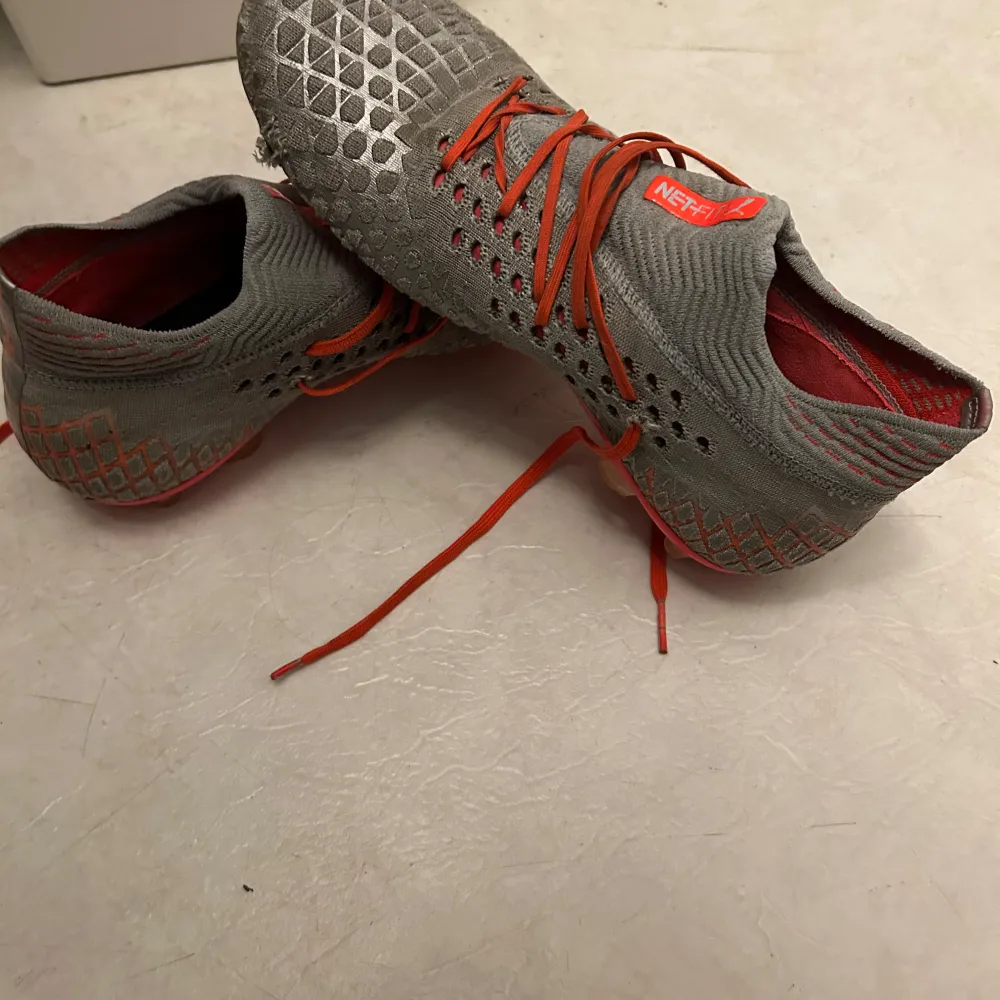 Säljer ett par gråa och orangea fotbollsskor från Puma i bra skick. Skorna har en snygg stickad design med orange snörning och en slitstark sula med dobbar för bra grepp på planen. Perfekta för fotbollsträning och matcher!. Kengät.