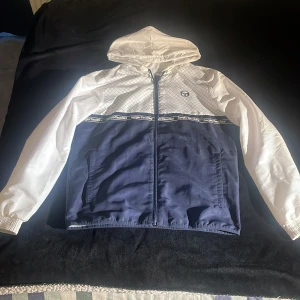 Vit och blå vindjacka från Sergio Tacchini - Säljer en snygg vit och blå vindjacka från Sergio Tacchini. Jackan har ett coolt rutmönster på den vita delen och en dragkedja framtill. Den har också en huva och elastiska ärmslut. Perfekt för vår och höst! Pris går alltid att förhandlas! Andvänd en handfull gånger Max!