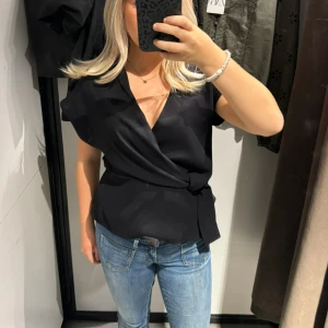Svart omlottblus - Snygg svart omlottblus med korta ärmar och knytning i midjan. Endast använd en gång!!! Strl m men passar s