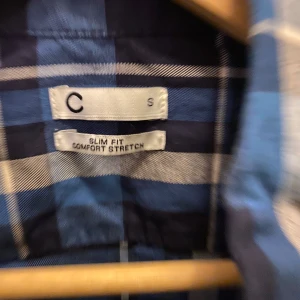 Blå och vit rutig skjorta - Säljer en snygg blå och vit rutig skjorta i slim fit med komfortstretch. Perfekt för både vardag och fest! Skjortan har långa ärmar och knappar hela vägen ner. Den är i bra skick och redo för nya äventyr. Passar perfekt till jeans eller chinos!