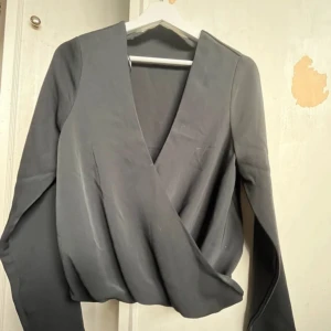 Svart omlottopp från BikBok - Snygg svart omlottopp från BikBok i storlek S. Toppen har långa ärmar och en stilren v-ringad design. Perfekt för både vardag och festliga tillfällen. Materialet känns mjukt och bekvämt mot huden. Helt använd men utan lappar då den ej passade.