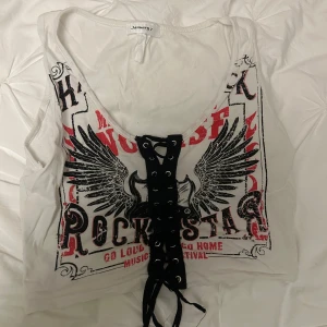 Vit rockig linne med snörning - Coolt vitt linne med rockigt tryck och snörning framtill. Designen har svarta vingar och text i rött och svart. Köpte på dolls kill för ca 300kr men aldrig använd så mitt pris 150! Så snygg!