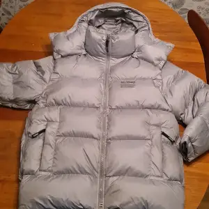 Säljer en snygg och varm silverfärgad pufferjacka från Axel Arigato. Jackan är tillverkad av 100% återvunnen polyester och dun, vilket gör den både miljövänlig och superbekväm. Perfekt för kalla vinterdagar med sin quiltade design och praktiska dragkedja. Ett måste för garderoben! ❄️