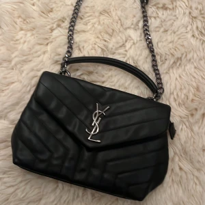 Svart ysl väska med kedja - Säljer en superfin svart quiltad väska från YSL. Den har en snygg kedja och ett elegant YSL-emblem på framsidan. Perfekt för både vardag och fest! 🖤