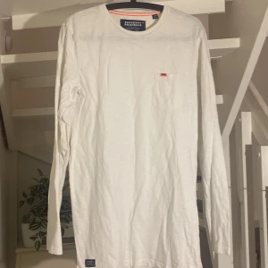 Vit långärmad tröja från Superdry - Säljer en stilren vit långärmad tröja från Superdry. Den har en liten bröstficka med en diskret Superdry-logga. Perfekt för en avslappnad look och passar till alla säsonger. Tröjan är i bra skick och gjord av mjukt bomullsmaterial.
