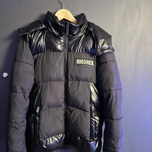 Svart pufferjacka från Hoodrich - Säljer en snygg svart pufferjacka från Hoodrich i storlek XS men passar S inte alls XS är själv 172 och den känns lite lång på mig men inget man tänker på så mycket. Jackan har en glansig finish med dragkedja och en stor huva. Perfekt för kalla dagar med sin varma vaddering. Stort Hoodrich-tryck på bröstet och ryggen ger en cool look. Passar perfekt för vintern och hösten!