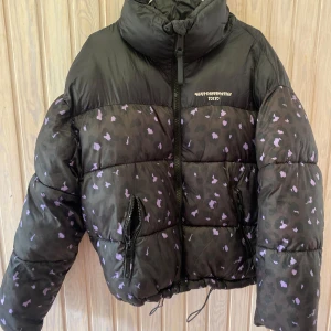 Svart pufferjacka från FB Sister - Säljer en cool svart pufferjacka från FB Sister med lila mönster. Jackan har en hög krage och dragkedja framtill. Perfekt för höst och vinter, håller dig varm och snygg! 🖤💜