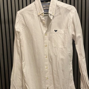 Vit skjorta från DeFacto - Säljer en stilren vit skjorta från DeFacto i storlek XS. Skjortan har en modern passform och är långärmad med snygga blå detaljer på insidan av manschetterna. Den är broderad med en liten blå detalj på bröstfickan. Perfekt för både vardag och fest! 💙