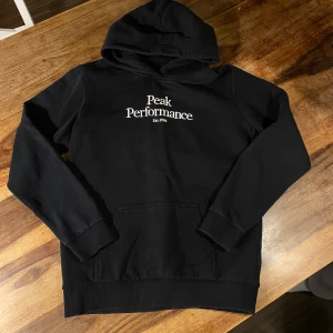 Svart hoodie från Peak Performance - Säljer en svart hoodie från Peak Performance i mycket bra skick. Den har en klassisk design med vit logga på bröstet och en stor ficka framtill. Perfekt för höst och vinter, och den är supermjuk och bekväm. Passar både till vardags och chillkvällar. 🖤