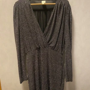 Glittrig svart mini klänning - Snygg svart klänning med glittrigt mönster, perfekt för festliga tillfällen. Den har en omlott-design med v-ringning och långa ärmar. Materialet är stretchigt och bekvämt, vilket ger en smickrande passform. Klänningen är i mycket bra skick och redo att glänsa på nästa fest! Inga prislappar kvar men oanvänd. 