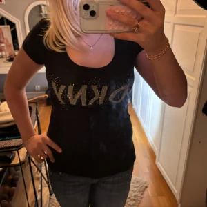 Svart t-shirt från DKNY - Snygg svart t-shirt från DKNY med glittrande logga framtill. Perfekt för en casual look eller att klä upp med accessoarer. T-shirten har en normal passform och korta ärmar. Ett måste i garderoben för alla som älskar stil och komfort!