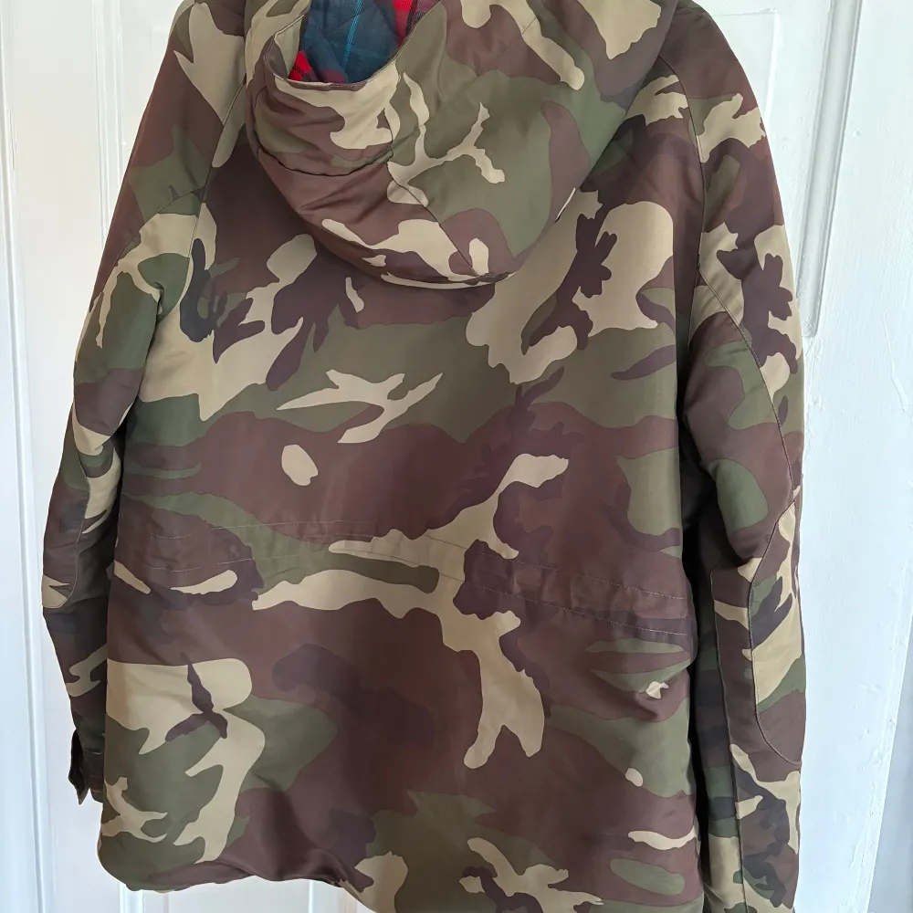 Säljer en cool camouflagejacka från These Glory Days i storlek medium. Jackan har ett klassiskt camouflagemönster och är fodrad med ett rött och blått rutigt tyg. Perfekt för höst och vår, med praktiska fickor och en skön huva. Passar både för vardag och äventyr!. Jackor.