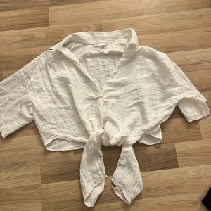 Vit knytblus i linne - Säljer en superfin vit blus i linne med knyt framtill. Perfekt för sommardagar med sin lätta och luftiga känsla. Blusen har korta ärmar och en avslappnad passform. Passar både till stranden och en dag på stan! Aldrig använd.