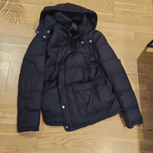 Mörkblå pufferjacka med huva från Boomerang  - Säljer en snygg mörkblå pufferjacka med huva. Jackan har långa ärmar och praktiska fickor framtill. Perfekt för kalla dagar och håller dig varm och bekväm. Passar både till vardags och lite finare tillfällen. Hör av dig vid intresse!