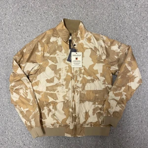 Tvåsidig jacka Woolrich - Säljer en snygg beige bomberjacka från Woolrich i nytt skick. Jackan har ett coolt camouflagemönster på ena sidan och är enfärgad på den andra, vilket gör den vändbar. Den har dragkedja framtill och är långärmad. Perfekt för höst och vår! Den passar mer som en S