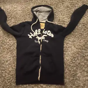 Säljer en snygg mörkblå hoodie från Hollister med vit text på framsidan. Den har en dragkedja och en mysig huva med vita snören. Perfekt för en avslappnad stil eller kyliga dagar. Passar både till vardags och chill.