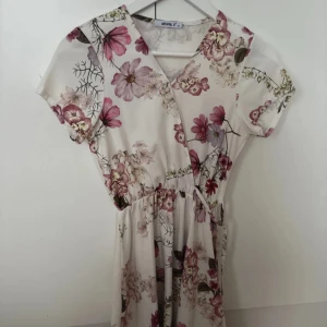 Blommig klänning - Säljer en superfin blommig klänning. Den är vit med rosa och lila blommor, har en omlott-design och korta ärmar. Klänningen är knappt använd, därav mycket bra skick🌸