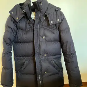 Säljer en navy dunjacka från Boomerang (modell Alexandra down jacket) i storlek XXS. Den är perfekt för kalla vinterdagar med sin tjocka vaddering och långa ärmar. Jackan har en praktisk huva och stängs med både dragkedja och knappar. Den är i bra skick och redo för nya äventyr!