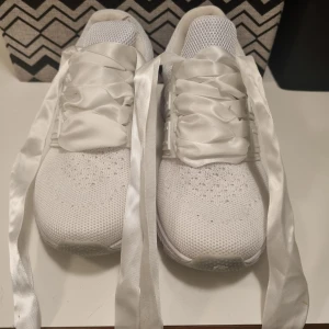 Vita sneakers med satinband - Säljer ett par vita sneakers i syntetmaterial med unika satinband som snörning. Skorna har en stilren design med en bekväm sula och är perfekta för både vardag och fest. De är i nytt skick och redo att användas direkt. 